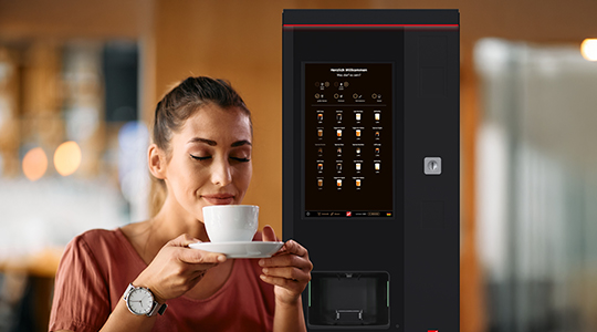 Eine Frau trinkt Kaffee aus einer Tasse, während sie vor einem SiLine HG TS27-Kaffeeautomat steht.